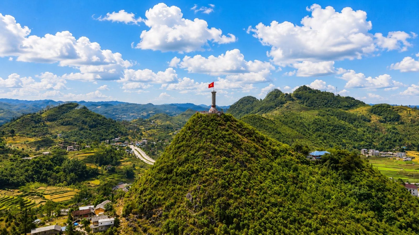 The Lũng Cú flag tower on a forested hilltop above a valley of terraced fields, blue sky with clouds