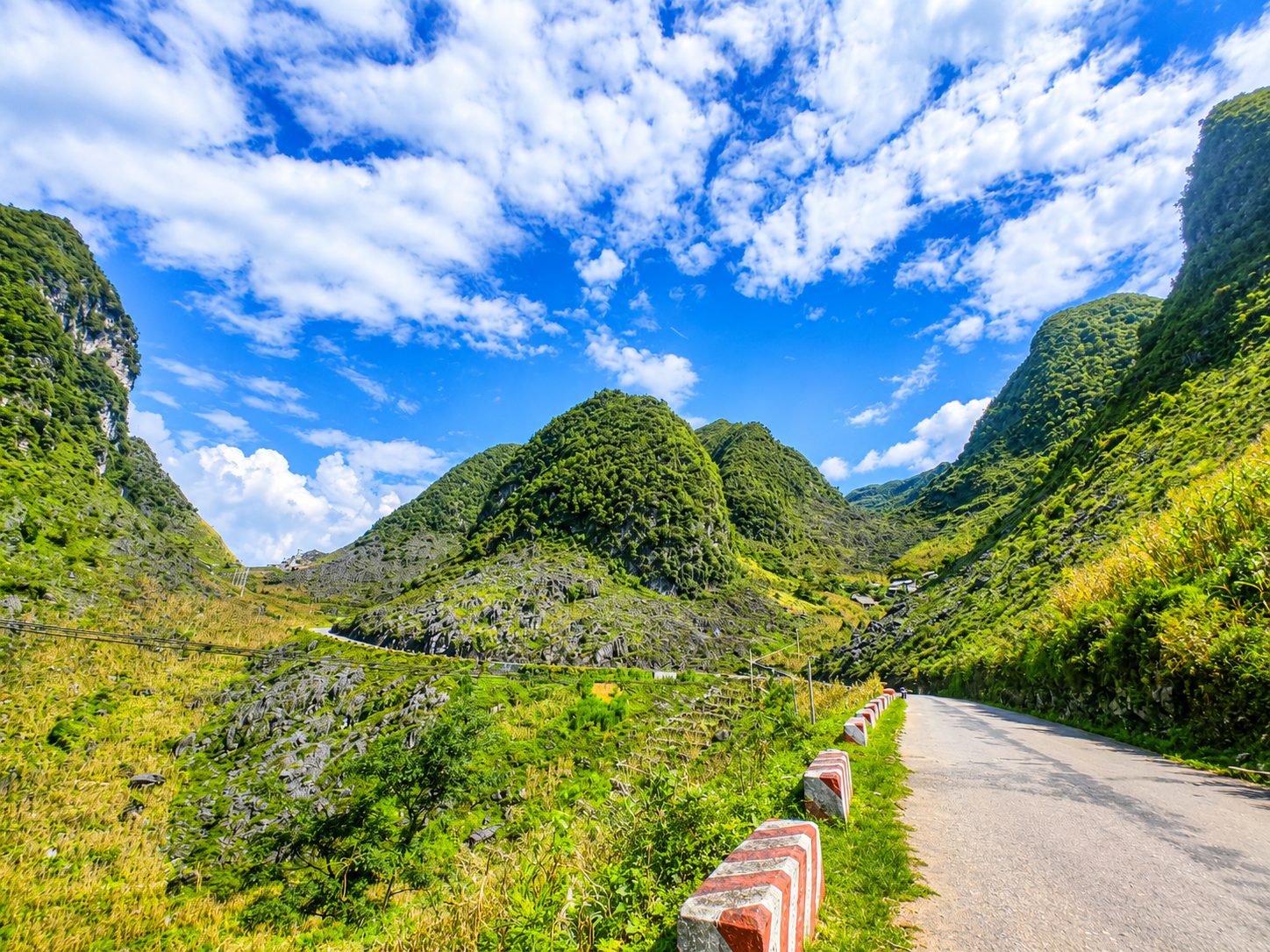 A quiet mountain road running past green karst peaks and rocky slopes under a bright blue sky