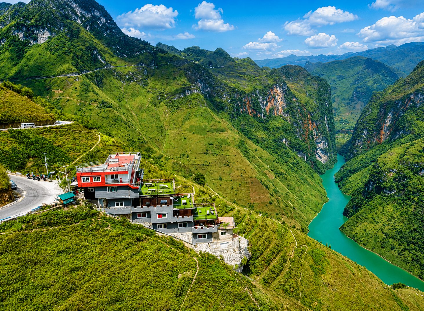 A red-roofed lookout building perched on a green mountainside above the turquoise Nho Quế River gorge at Mã Pí Lèng