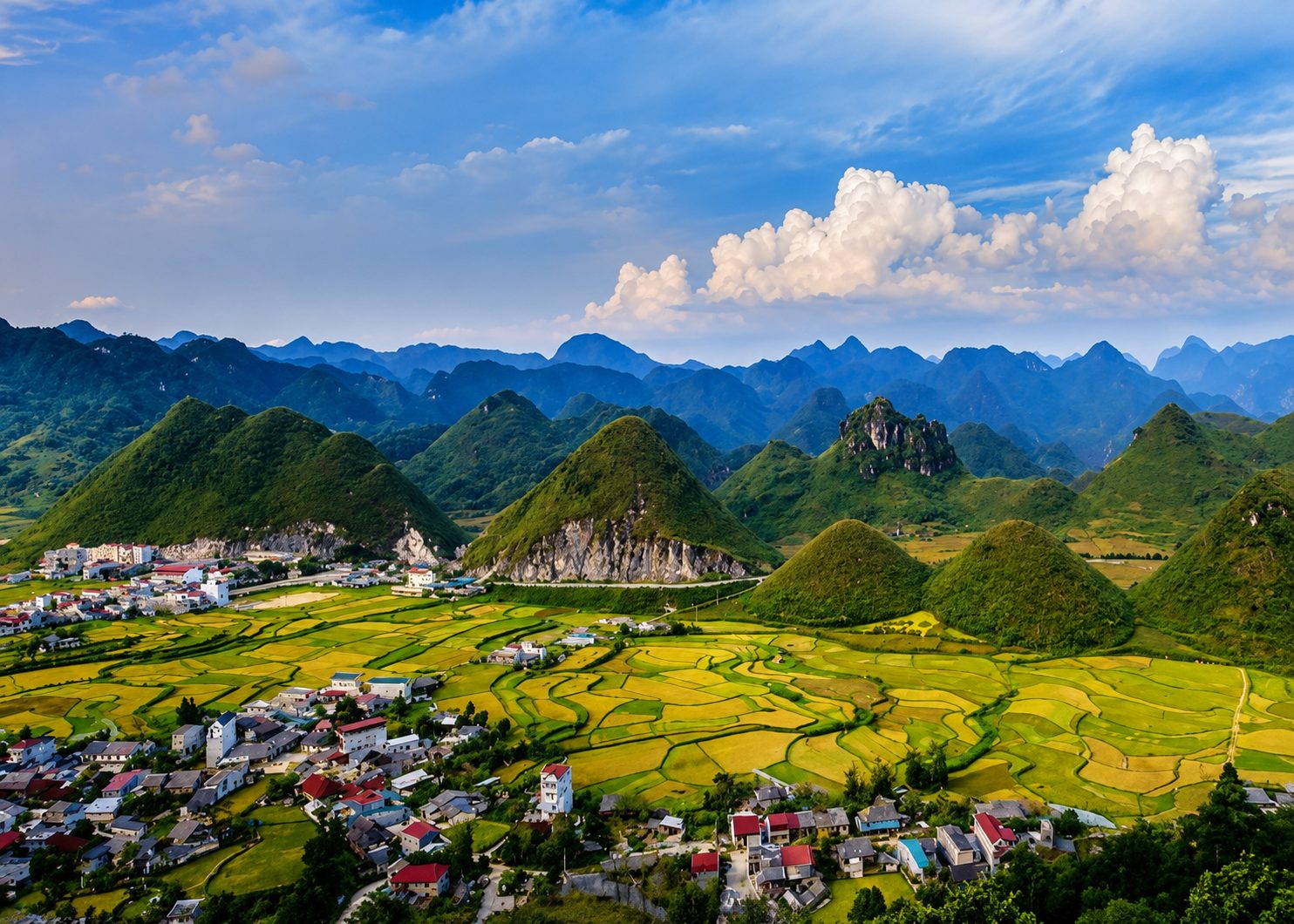 Cone-shaped karst hills rising behind golden rice fields and a small town in Quản Bạ, under white clouds