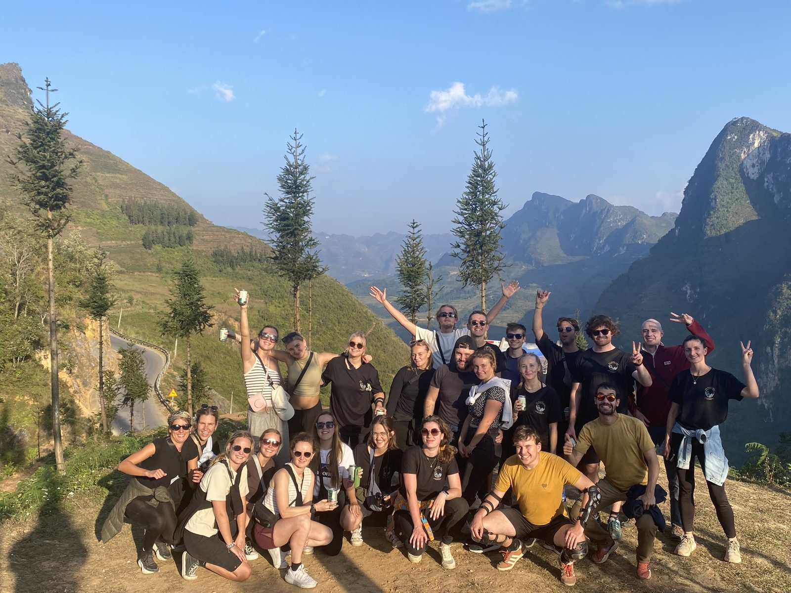 A big group of travellers cheering at a mountain viewpoint with pine trees and karst peaks behind