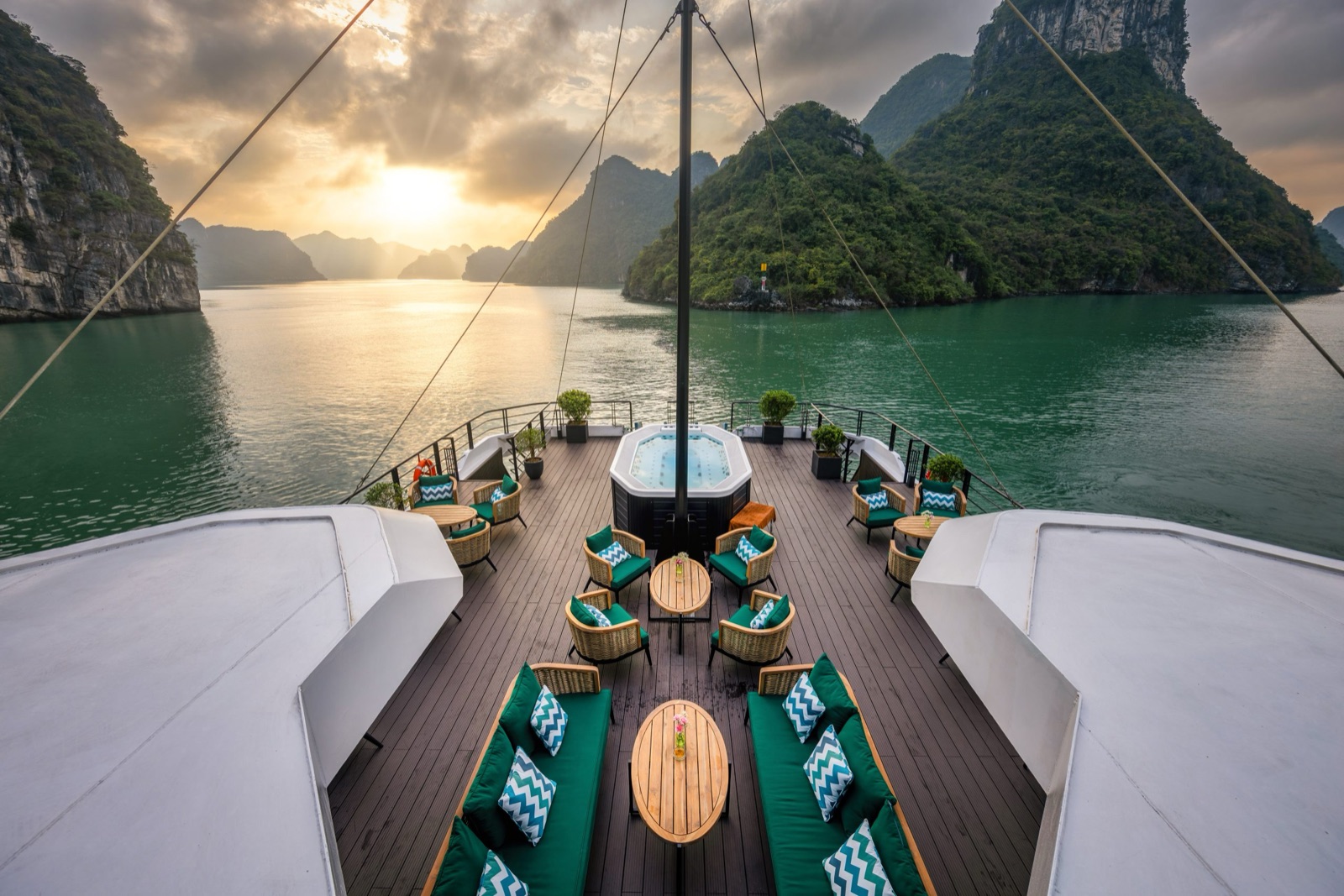 The catamaran’s bow deck with green sofas and a jacuzzi, the sun setting between karst peaks
