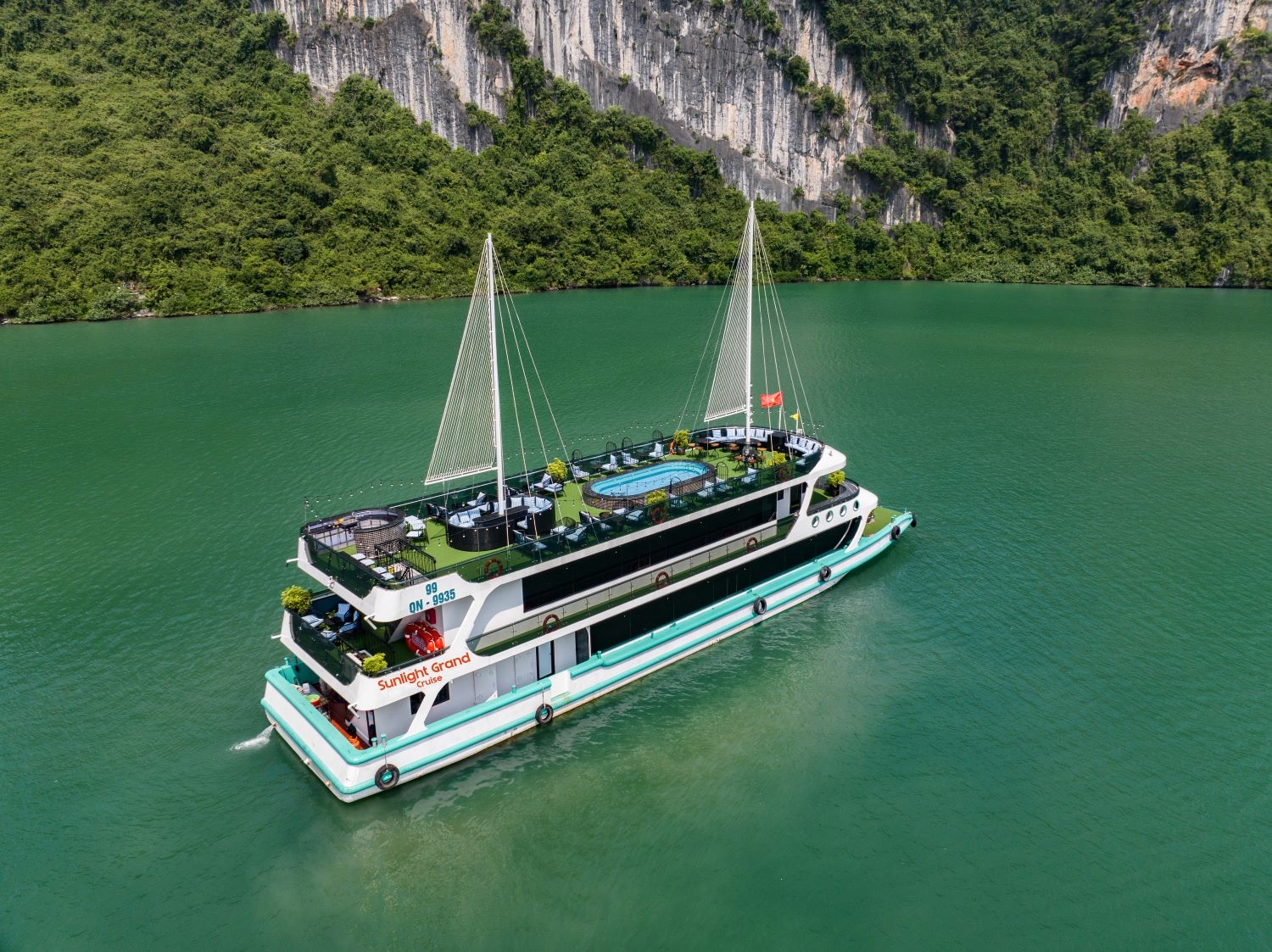Aerial view of the two-masted Sunlight Grand boat on green water below a limestone cliff, a pool on its top deck