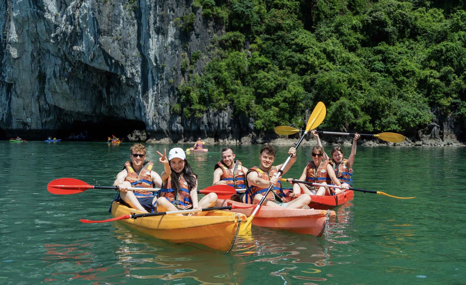 A group of friends in life jackets raising their paddles in kayaks below a jungle-covered cliff