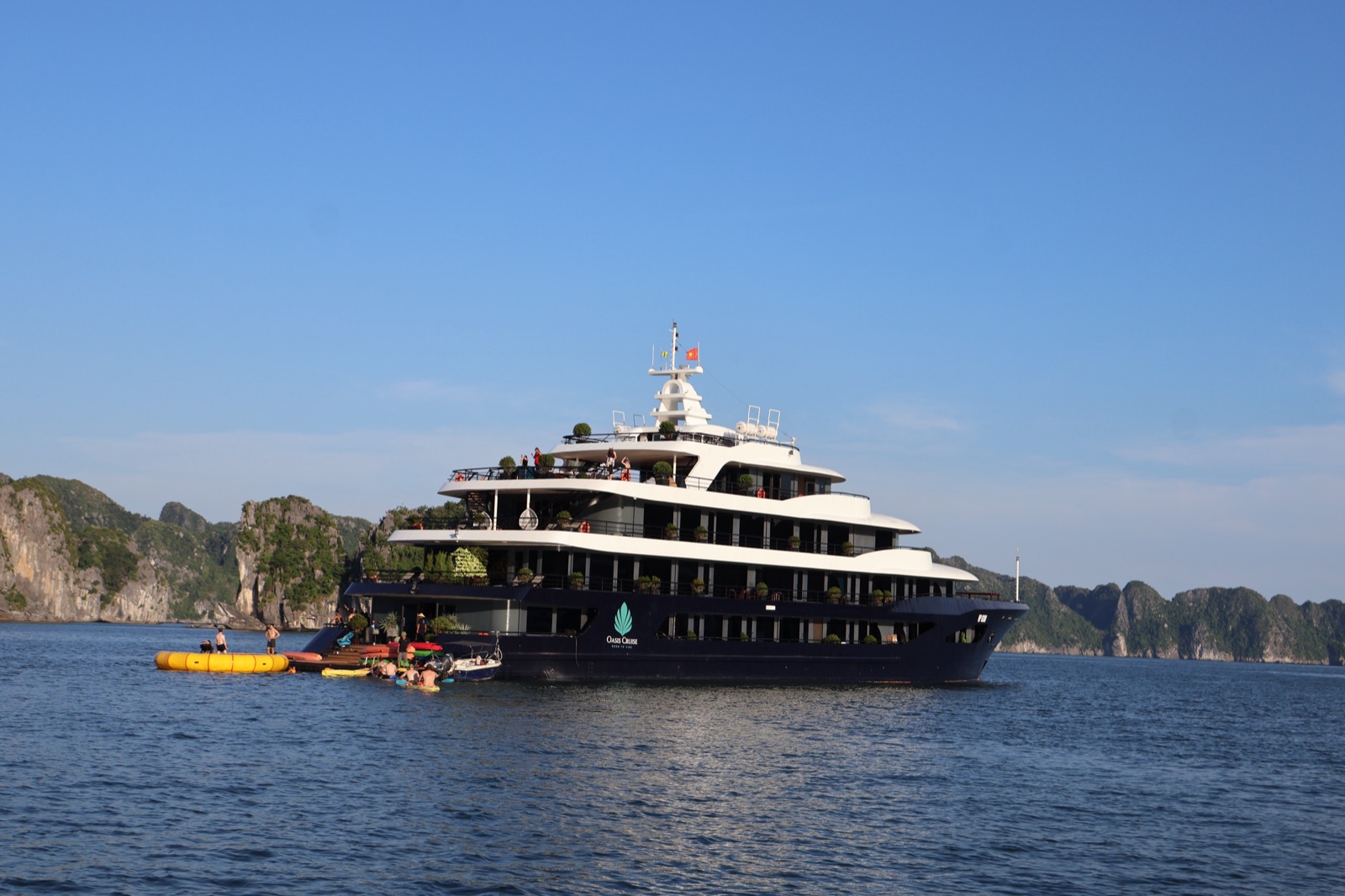 The dark-hulled Oasis Bay party boat at anchor in Hạ Long Bay, a yellow water trampoline beside it
