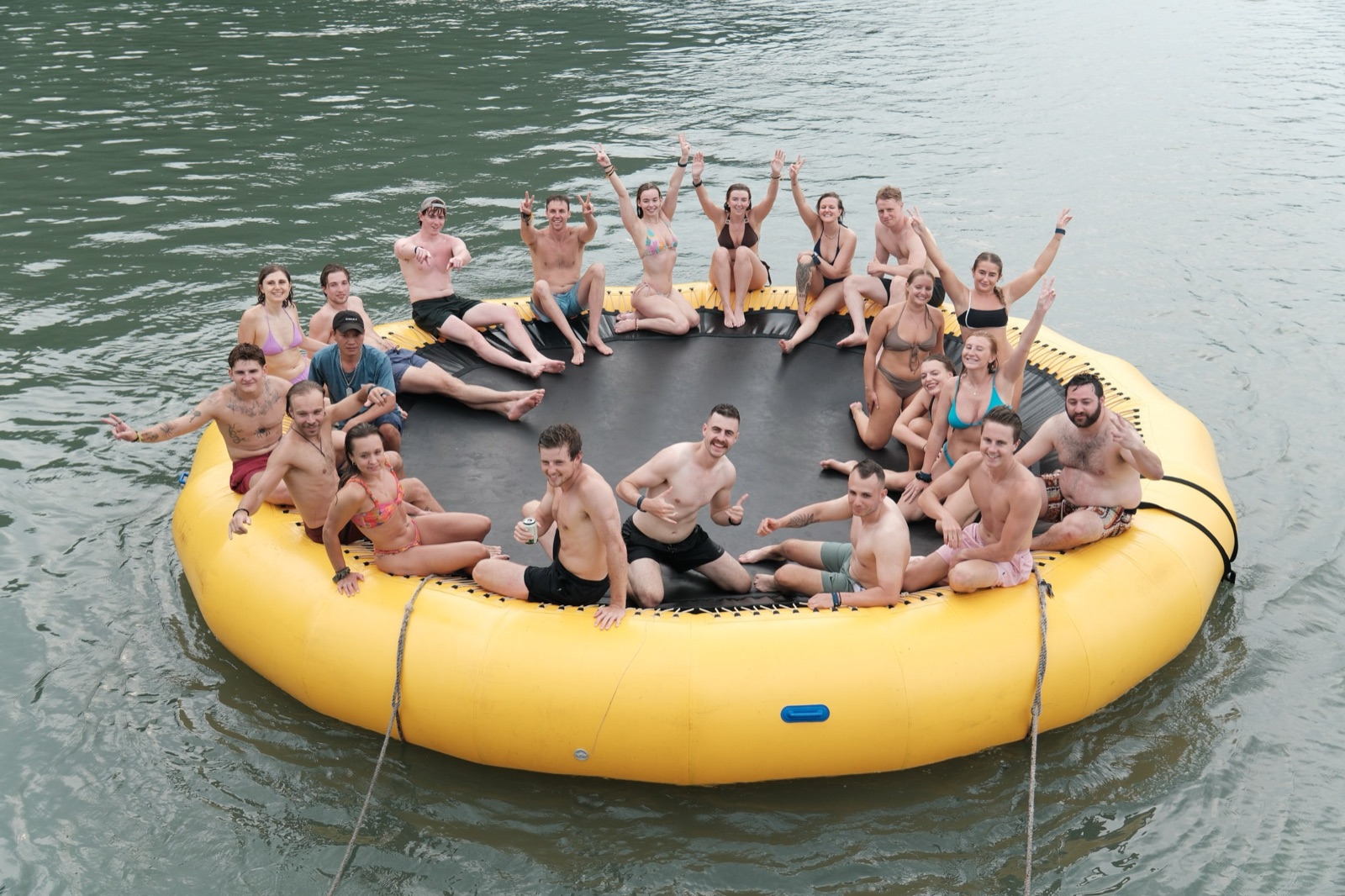 A crowd of guests in swimwear waving from a yellow water trampoline in the bay