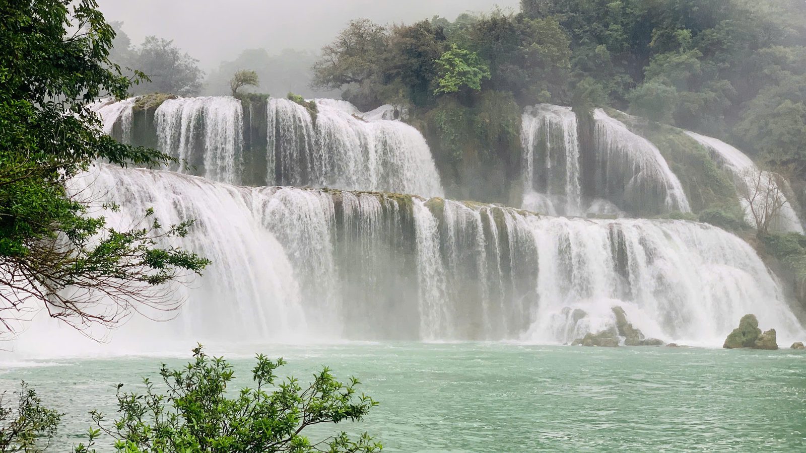 Bản Giốc waterfall pouring over wide rocky tiers into a green river, mist rising