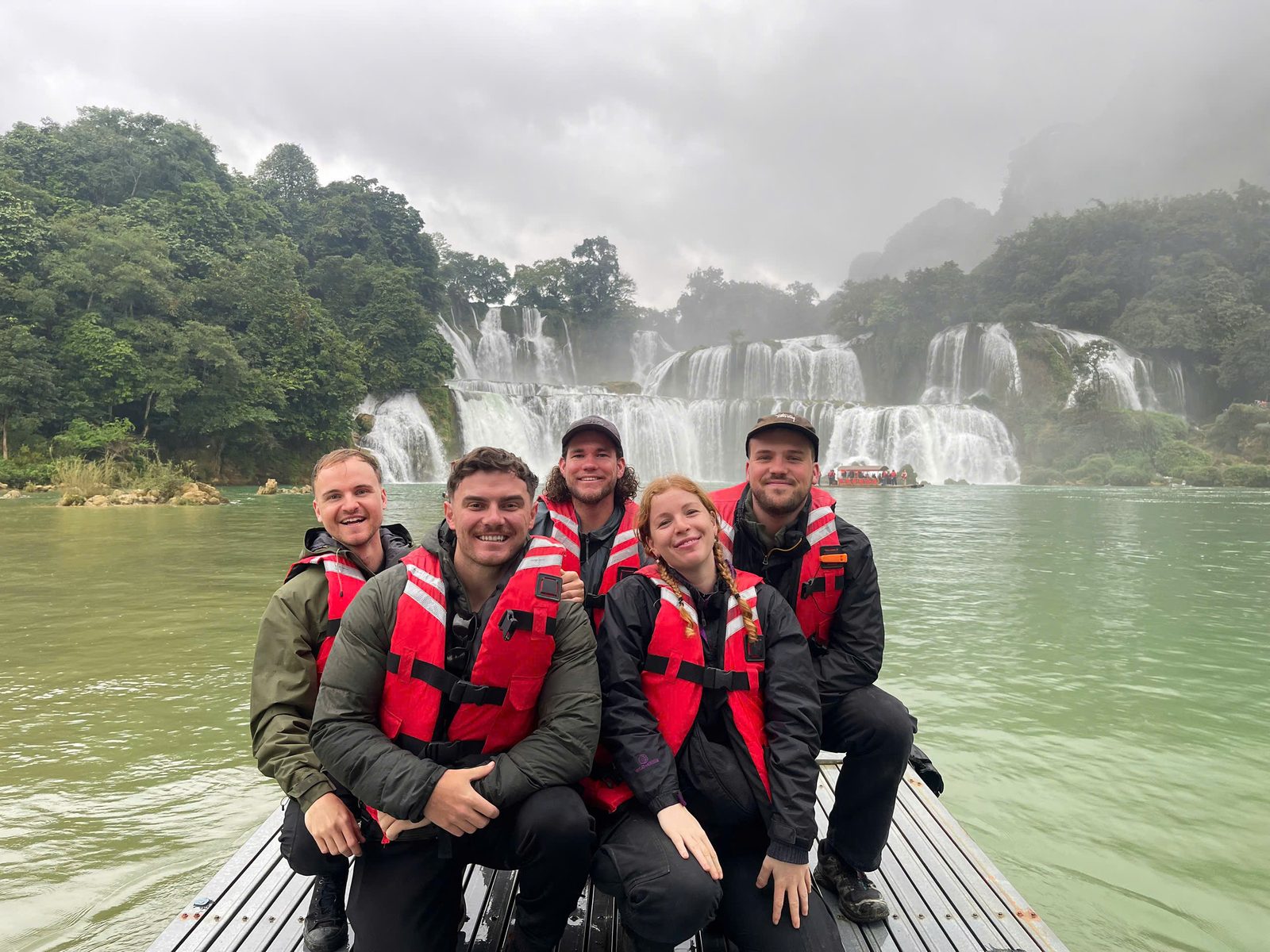 A group in life jackets on a raft in front of Bản Giốc waterfall on a misty day