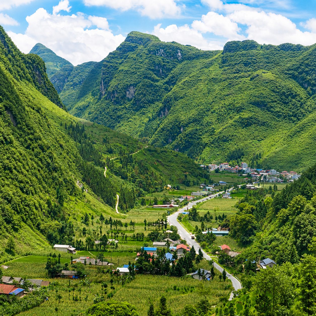 A green valley with a village and a road between steep mountains, white clouds above