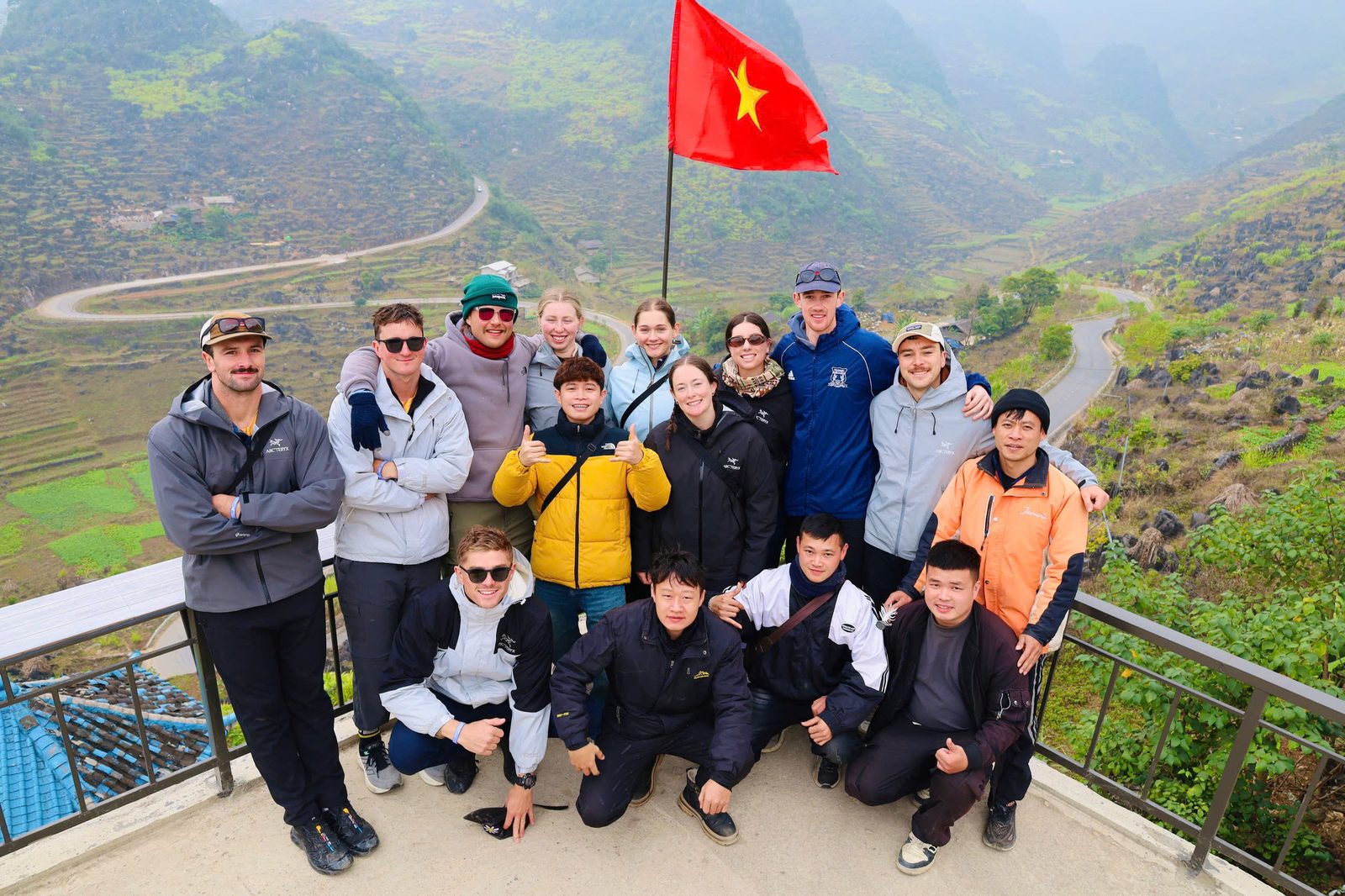 A tour group with their local crew at a viewpoint by a flag, a pass winding below