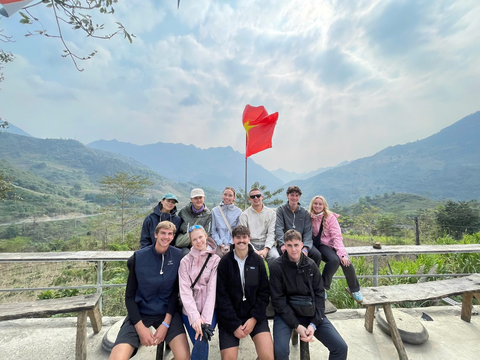 A group of travellers on wooden benches at a hill viewpoint, a red flag behind them